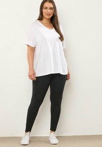 Losse, witte V-hals t-shirt met opgerolde korte mouwen, gecombineerd met zwarte leggings en witte sneakers, tegen een neutrale achtergrond.