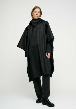 Svart vattentät poncho med huva, med en lös och oversized design samt slät yta. Återfinns tillsammans med mörkgrå byxor och svarta stövlar.
