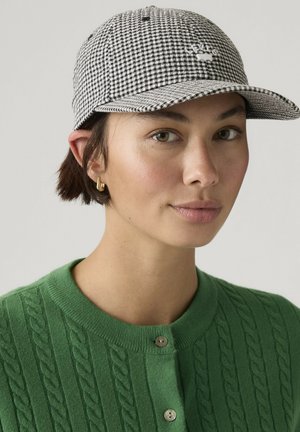 Jonge vrouw met kort donker haar draagt gouden creooloorbel, groene kabelgebreide cardigan en zwart-witte geruite baseballpet met logo.