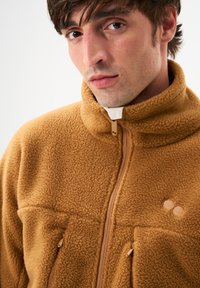 Fleecejacke in warmem Braun mit hohem Kragen, Reißverschluss, zwei Fronttaschen und strukturierter Oberfläche. Zarte Markenakzente sind vorhanden.