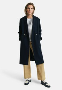 Marineblauwe wollen jas met gouden knopen, gedragen over een gestreept shirt, gecombineerd met tan kleurige wijde broekspijpen en zwarte loafers met witte accenten.