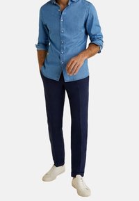 Camisa de mezclilla abotonada en azul claro, combinada con pantalones chinos azul marino y zapatillas blancas. La camisa presenta un cuello clásico y mangas largas.