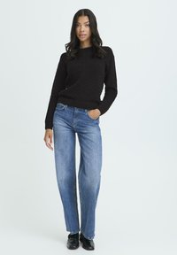 Kvinde står med den ene hånd i lommen, iført en sort tekstureret sweater, blå lige ben jeans og sorte sko, foran en ensfarvet hvid baggrund.