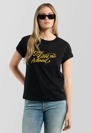 Junge Frau trägt schwarze Sonnenbrille und ein schwarzes T-Shirt mit gelbem Schriftzug "Gas Fuel the Mood", kombiniert mit hellblauen Jeans.