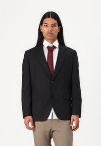 Must blouse'ga ja beežide pükste üle kantud must täpselt õmmeldud bleiser, millel on kaks esi-taskut, nööbiga sulgemine ja sälk-lapell.