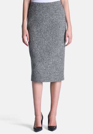 Femme portant une jupe crayon grise taille haute, longueur genou, avec des chaussures à talons noirs, debout devant un fond uni.