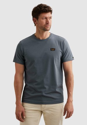 Man draagt een aansluitend grijs T-shirt met korte mouwen met een klein logopatch op de borst en lichtbeige broek.