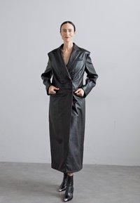 Schwarzer Leder-Langmantel mit doppelter Knopfleiste, scharfem Kragen, Seitentaschen und glatter Textur. Trägt man über schwarzen hochhackigen Stiefeln.