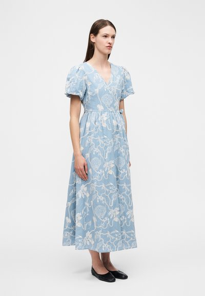 Žena v světle modrých midi šatech s bílou výšivkou s mořským motivem, nabíranými rukávy, výstřihem do V a černými balerínami, stojící na bílém pozadí.