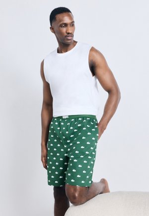 Hombre con camiseta sin mangas blanca y pantalones cortos verdes con estampado, apoyando una rodilla sobre una superficie beige texturizada, mirando hacia un lado.