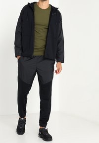 Musta vetoketjullinen takki hupulla tummanvihreän t-paidan yli, yhdistetty mustiin housuihin, joissa on mattapintaa ja kiiltävää tekstuuria. Urheilukengät.