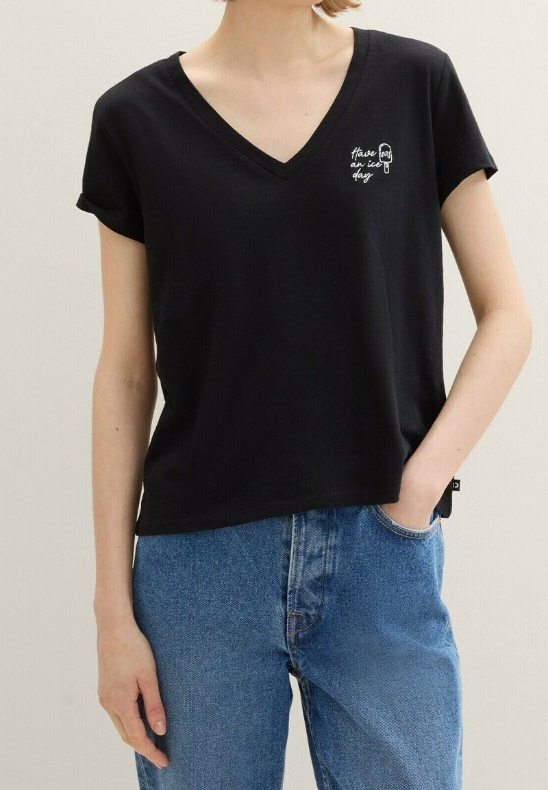 Personne portant un T-shirt noir en V avec "Have an ice day" et un graphique de glace, assorti d'un jean bleu, main dans la poche.