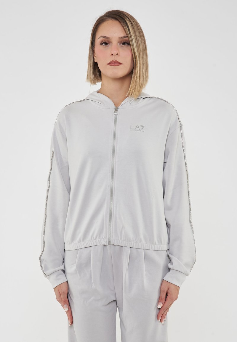 Giacca grigia con zip e cappuccio, con accenti argentati lungo le maniche, tessuto morbido e vita aderente. Logo sul petto.
