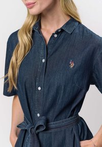 Tmavě modré džínové šaty košilového střihu s krátkými rukávy, zapínáním na knoflíky vpředu, límečkem, zavazováním v pase a vyšívaným logem na hrudi. Hladká textura.