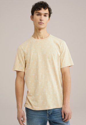 Tricou bej deschis cu un model geometric alb, decolteu rotund și mâneci scurte. Materialul pare moale și casual. Purtați-l cu blugi albaștri.