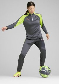 Sivi sportski trenirka s neonskim žutim naglascima, s visokim ovratnikom i zatvaračem, uparena s neonskim fudbalskim kopačkama i šarenom crnom i zelenom fudbalskom loptom.