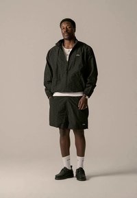 Uomo che indossa una giacca nera con motivo e zip abbinata a pantaloni corti, calze bianche e scarpe nere, in piedi contro uno sfondo beige uniforme.