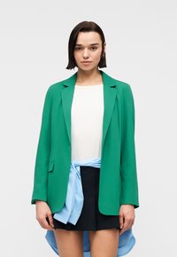 Femme aux cheveux courts et foncés, portant un blazer vert, un haut blanc, une jupe noire et une chemise bleu clair nouée autour de la taille, face à l'avant.