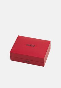 Boîte-cadeau rectangulaire rouge à texture lisse, avec un logo noir "HUGO" affiché de manière bien visible sur le couvercle.