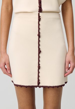 Gonna crema con un taglio dritto, caratterizzata da un bordo testurizzato bordeaux e un orlo ondulato. Decorata con un accento centrale in crochet bordeaux.