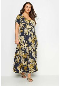 Abito lungo con sfondo blu navy e motivo floreale giallo, con maniche corte e scollatura a V profonda, abbinato a sandali dorati.