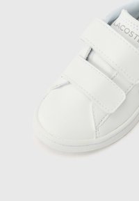 Λευκό δερμάτινο αθλητικό παπούτσι με στρογγυλεμένη μύτη, δύο ρυθμιζόμενους ιμάντες με Velcro και διακριτικές ραφές. Το λογότυπο της Lacoste εμφανίζεται στον ιμάντα.