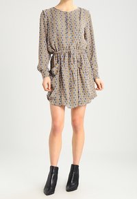 Sukienka z długim rękawem w kwiatowy wzór w diamenty w kolorze żółtym, niebieskim i białym. Elastyczna talia, luźny krój i falbaniasty dół. Noszona z czarnymi botkami za kostkę.