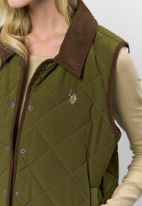 Pikowana oliwkowa kamizelka z brązowym kołnierzem z sztruksu i zatrzaskami. Posiada haftowany wzór oraz boczne kieszenie. Teksturowana powierzchnia.