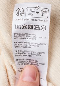 Etichetta di cura realizzata in tessuto, prevalentemente bianca con testo nero. Include istruzioni per il lavaggio in più lingue e simboli di cura.