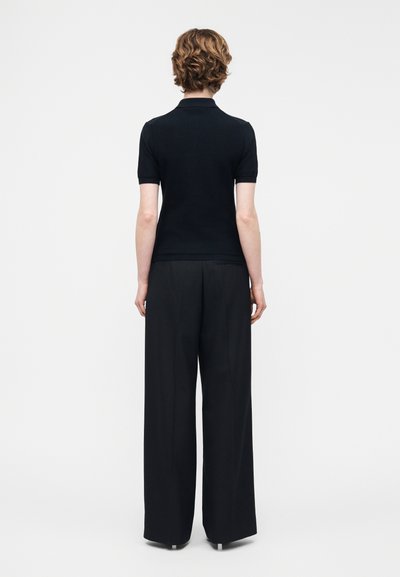 Άτομο με κοντά σγουρά μαλλιά, που φοράει μαύρο πουκάμισο πόλο με κοντά μανίκια και μαύρο παντελόνι με ευρύ πόδι, στέκεται με την πλάτη προς τα πίσω σε λευκό φόντο.