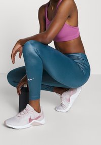 Leggings teal taille haute avec une finition brillante, assortis à un soutien-gorge de sport rose et des baskets Nike blanches et violettes, accroupie sur une bouteille d'eau.