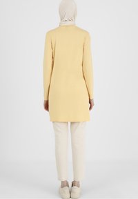 Nainen, jolla on päällään beige huivi, keltainen pitkähiha-tunika, vaaleanbeiget housut ja vaaleanbeiget lenkkarit, seisoo kasvot poispäin valkoista taustaa vasten.