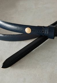 Sötétkék bőr öv sima felülettel, arany fém csattal és vékony, keskenyedő dizájnnal. A széleken varrási részletek találhatóak.