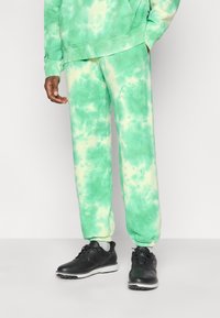 Το πράσινο φούτερ με σχέδιο tie-dye και οι αντίστοιχες φόρμες διαθέτουν απαλή υφή. Μαύρα παπούτσια με λευκή σόλα και κορδόνια ολοκληρώνουν την εμφάνιση.