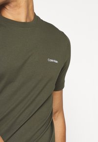Olivgrünes T-Shirt aus Baumwolle mit Rundhalsausschnitt und kurzen Ärmeln. Das Calvin Klein-Logo ist in Weiß auf der Brust gestickt.