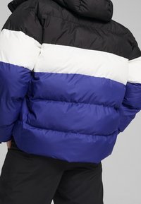 Pružná bunda se svislými černými, bílými a modrými pruhy. Vyrobena z lesklého prošívaného materiálu s kapucí a hranatým střihem.