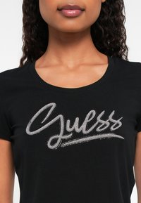 Kobieta w czarnej koszulce z srebrnym, błyszczącym logo "Guess" umieszczonym na środku klatki piersiowej.