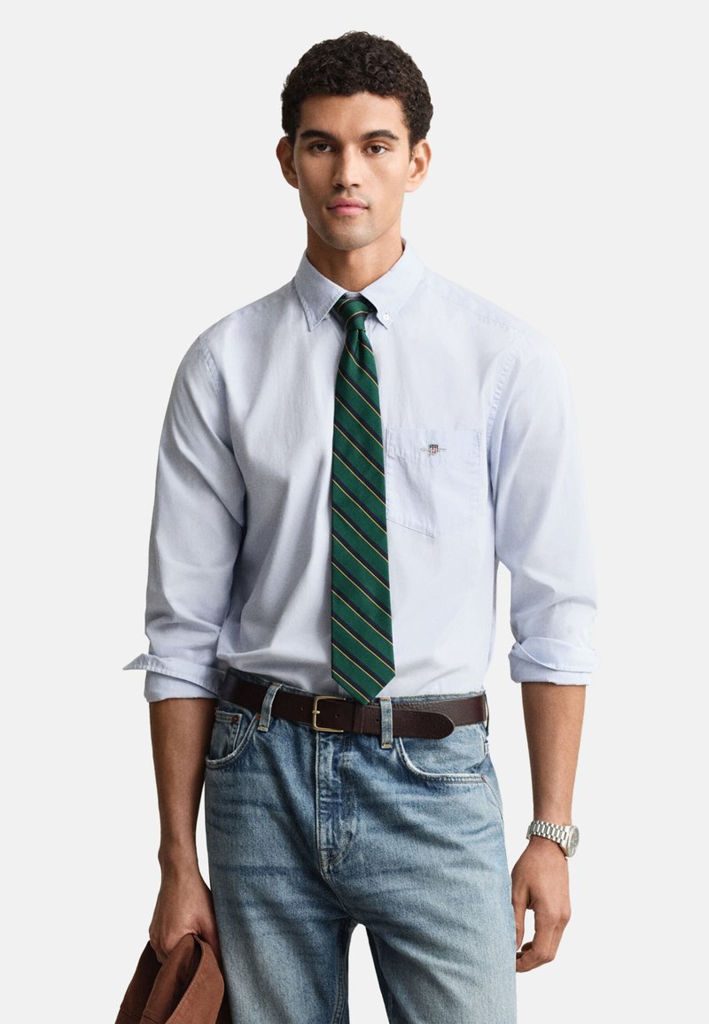 Lichtblauw overhemd met opgerolde mouwen, groene en gouden stropdas met strepen, bruin leren riem en vervaagde blauwe spijkerbroek, met een bruine jas in de hand.