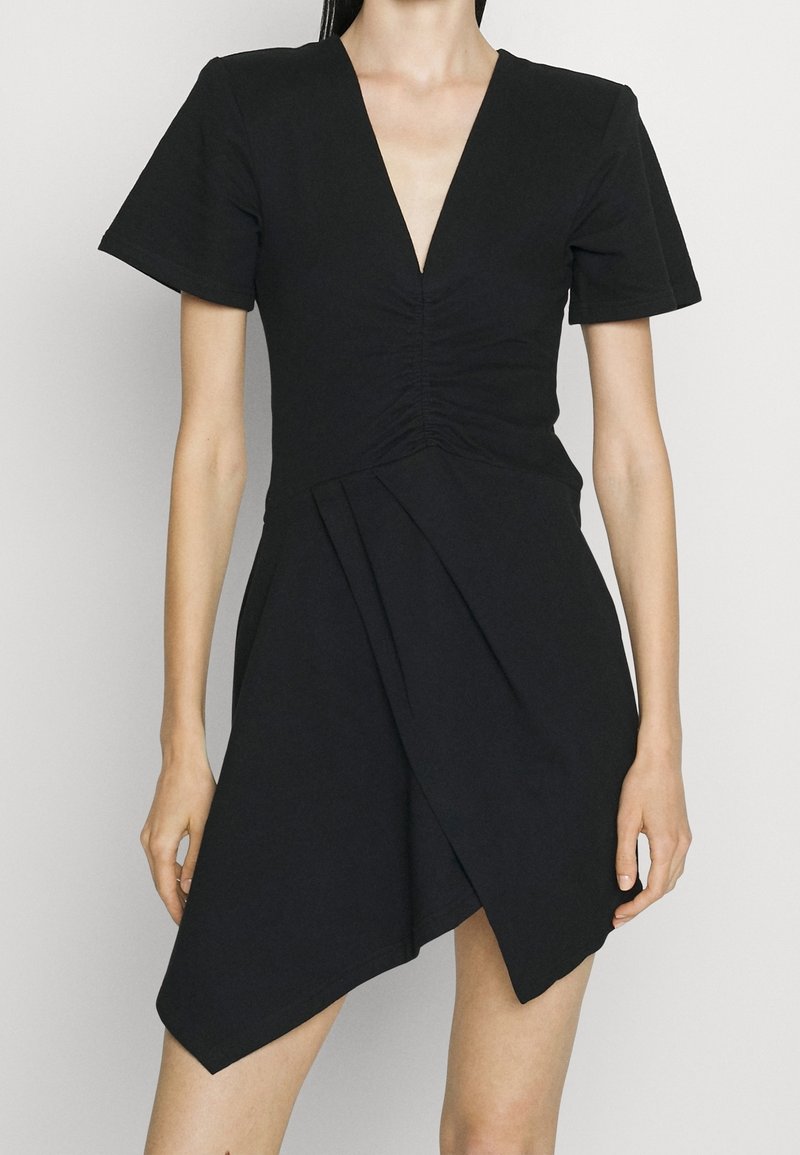Femme portant une robe noire courte avec des manches courtes, un profond décolleté en V, un devant froncé et un ourlet asymétrique.