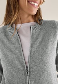 Cardigan grigio realizzato in tessuto morbido, con collo a coste e chiusura con zip dotata di un cursore tondo. Indossato sopra una camicia bianca.