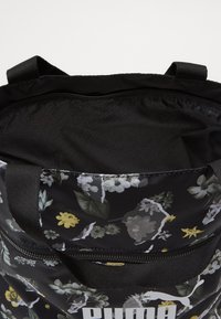 Borsa nera con stampa floreale che presenta motivi bianchi, gialli e verdi. Include una chiusura con zip e una fodera nera imbottita con branding discreto.