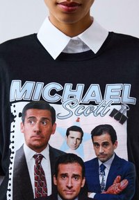 Čierne tričko s viacerými potlačami muža, s textom "MICHAEL SCOTT" nad ním, na štruktúrovanom pozadí pripomínajúcom papier na zápis.