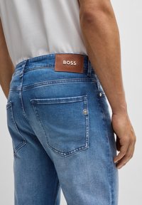 Niebieskie jeansy z denimu z brązową skórzaną naszywką z logo "BOSS". Posiadają przeszywane kieszenie oraz dopasowany, wąski krój. Gładka faktura.