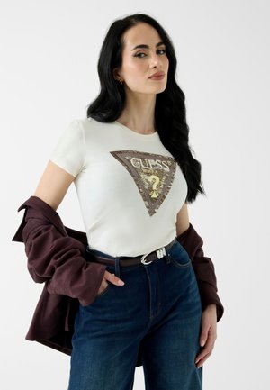 Femme aux longs cheveux foncés portant un t-shirt blanc avec le logo GUESS, un jean bleu foncé et une veste bordeaux drapée sur ses bras.