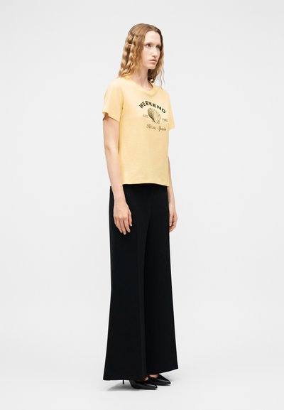 Femme aux cheveux ondulés portant un T-shirt jaune à motifs graphiques et un pantalon noir large taille haute, debout devant un fond blanc uni.