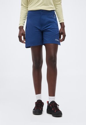 Osoba nosící modré outdoorové šortky se zipovými kapsami, béžové tričko s dlouhým rukávem, bílé ponožky a černé turistické boty s červenými tkaničkami.