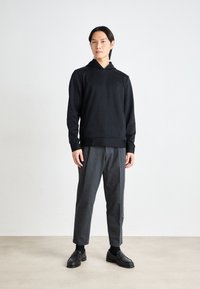 Black ribbed sweatshirt with hood and subtle logo, paired with gray pinstriped trousers and black loafers, worn on a plain background.