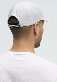 White baseball cap with a curved brim, featuring a striped adjustable strap at the back. Plain white cotton shirt visible in the foreground.