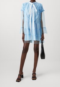 Ljusblå, genomskinlig klänning med volangdetaljer, knappstängning fram och knytkrage. Har långa, transparenta ärmar med prickar.