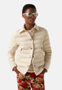 Světle béžová prošívaná bunda s límcem, vodorovným prošitím, dvěma kapsami na zip, doplněná kalhotami s květinovým vzorem a slunečními brýlemi.